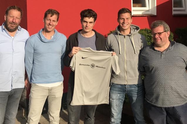 Gruppenbild vor einem roten Vereinsgeb&auml;ude von Borussia M&uuml;nster mit f&uuml;nf M&auml;nnern, die ein Trikot halten.