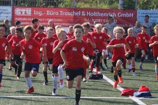 Gruppentraining von Kindern in roten Trikots, die auf einem Kunstrasenplatz sprinten. Zuschauer im Hintergrund.