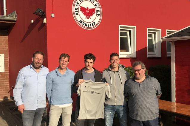 Gruppenbild vor einem roten Vereinsgeb&auml;ude von Borussia M&uuml;nster mit f&uuml;nf M&auml;nnern, die ein Trikot halten.