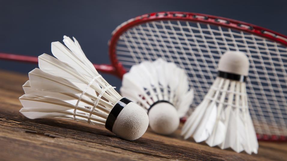 Drei Federbälle liegen auf einem Holztisch, mit einem Badmintonschläger im Hintergrund.