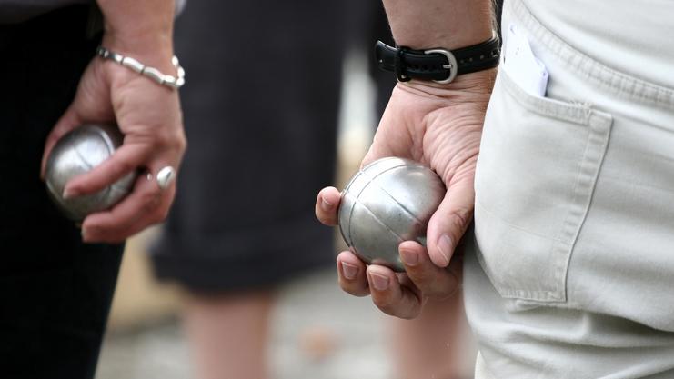 Zwei Personen halten silberne Kugeln in der Hand, während sie auf einem Platz an einem Boule-Spiel teilnehmen.