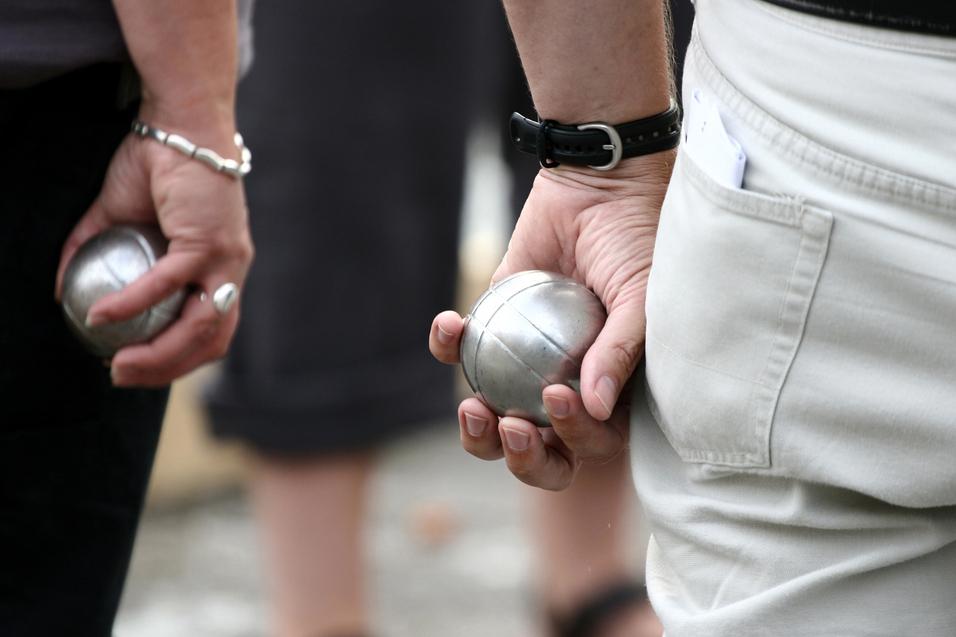 Zwei Personen halten silberne Kugeln in der Hand, während sie auf einem Platz an einem Boule-Spiel teilnehmen.