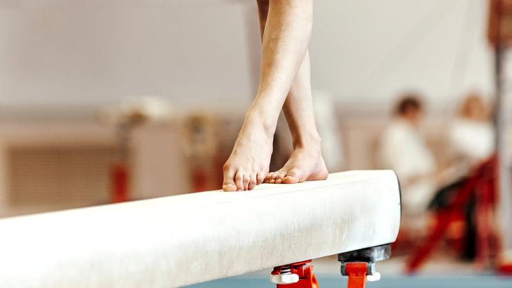 Fuß einer Turnerin auf dem Schwebebalken in einer Trainingshalle, im Hintergrund unscharfe Zuschauer.